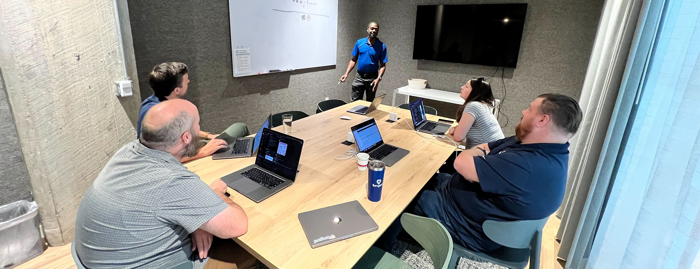 3 men and one woman sitting at a conference room table with laptops open on the table. Their attention is directed at a man standing in front of a whiteboard speaking.